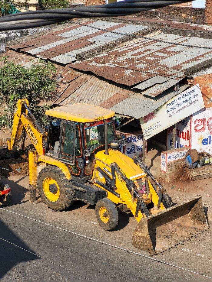Yellow backhoe loader parked near a rustic building with corrugated metal roof on a sunny day.