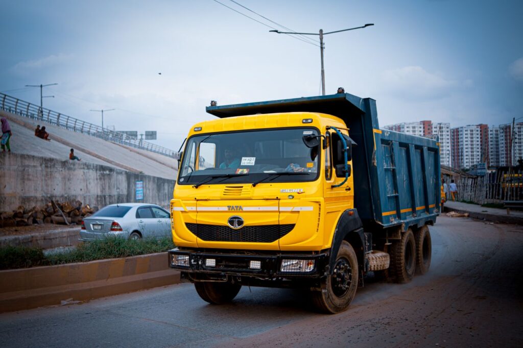 A yellow TATA dump truck traveling along an urban road in Dhaka, Bangladesh with urban housing in the background.